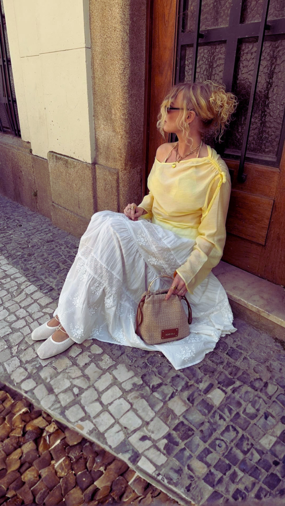 A blonde woman with her hair in a curly updo sits outside on a cobblestone path next to a rustic wooden door. She wears a relaxed, long-sleeved butter yellow top, a tiered white eyelet maxi skirt, and woven white mesh flats. She rests her hand on a small, structured beige woven straw bucket bag with a round gold top handle and brown leather trim.