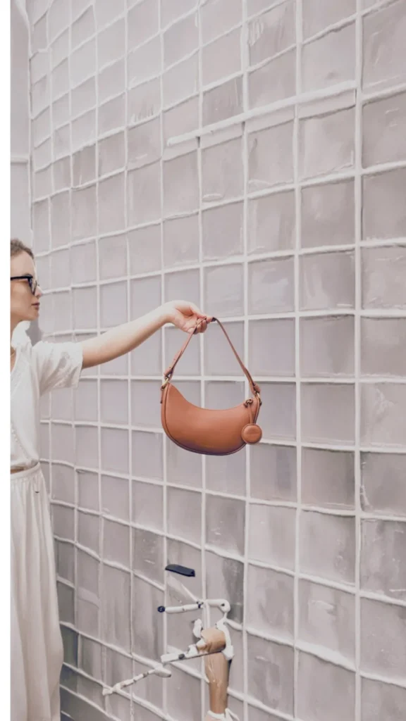 A woman stands in profile holding out a smooth, tan leather crescent-shaped shoulder bag by its strap. She wears a relaxed, cream-colored dress or top and dark-framed glasses. She is posing in front of a modern wall constructed from translucent square glass blocks, which diffuse the light softly across the scene.