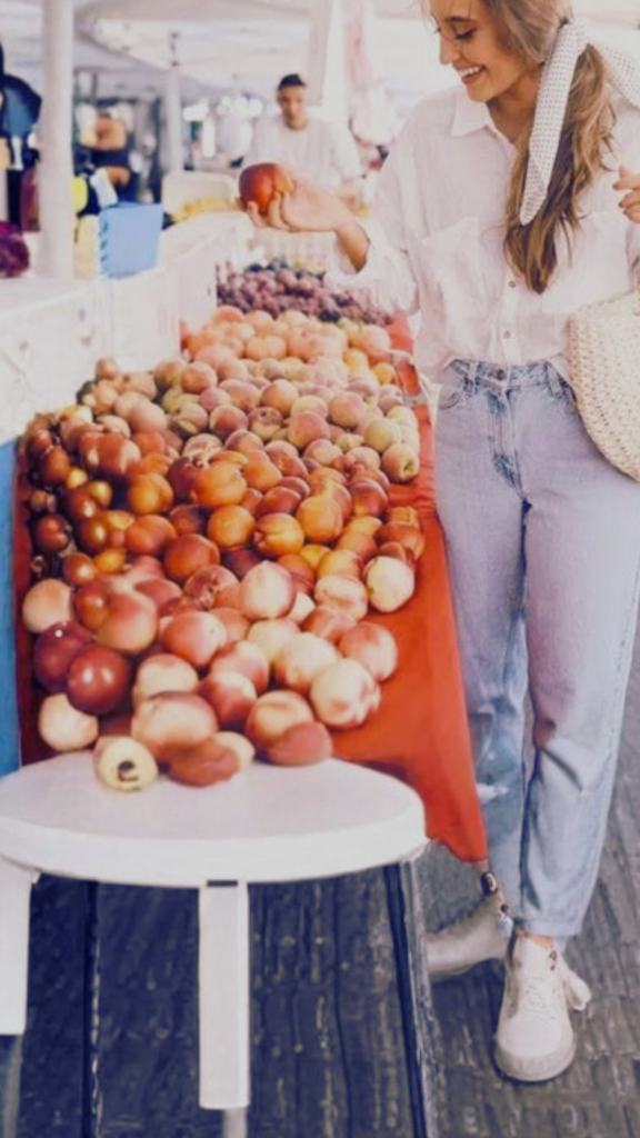 A young woman smiles as she stands at an outdoor farmer's market stall filled with fresh peaches. She wears a casual, breezy white button-down shirt, light wash denim jeans, white sneakers, and a white polka-dot scarf tied in her hair. She carries a woven straw tote bag under her arm as she reaches out to pick up a piece of fruit.