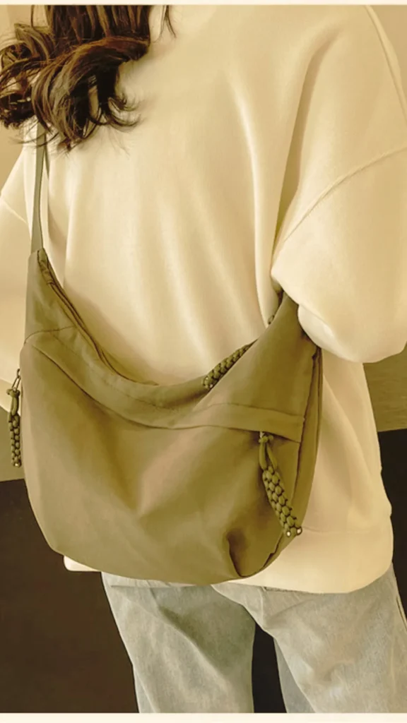 A close-up, back-angle view of a person with long, wavy brown hair wearing an oversized cream-colored sweatshirt and light-wash blue jeans. They are carrying a slouchy, crescent-shaped nylon sling bag in an olive green color across their back. The bag features a long shoulder strap and zipper pulls with thick, braided knotted details.