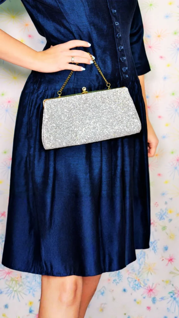 A close-up shot of a woman modeling an elegant evening outfit, featuring a shimmering silver glitter clutch bag. The structured handbag has a classic clasp closure and a fine gold chain handle that the woman is holding with her hand resting on her waist. She is wearing a rich, dark blue silk or satin dress with slight texturing and pleated detailing on the skirt, set against a light background with subtle starburst patterns.