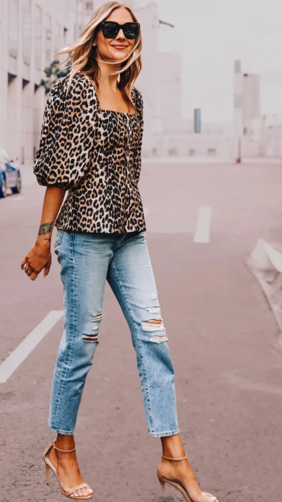 A woman smiling while walking down a city street, showcasing a chic, smart-casual outfit. She wears a square-neck, puff-sleeved top with a bold leopard print pattern, paired with light-wash blue jeans that feature distressed rips at the knees. Her look is completed with large black sunglasses, delicate gold rings, a tattoo on her wrist, and elegant nude strappy high-heeled sandals.