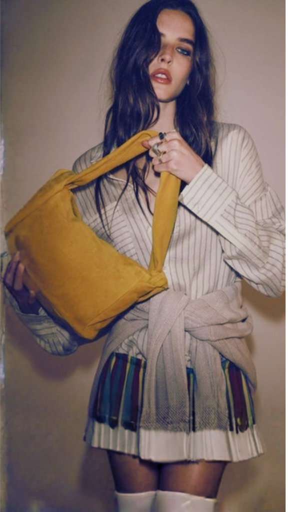 A model with long, wavy brown hair poses in a striped button-down shirt and a pleated mini skirt with colorful paneling. A neutral-toned knit sweater is tied around her waist. She holds up a medium-sized, slouchy mustard yellow suede shoulder bag with a thick knot detail on the strap, pulling focus to its vibrant, soft texture.
