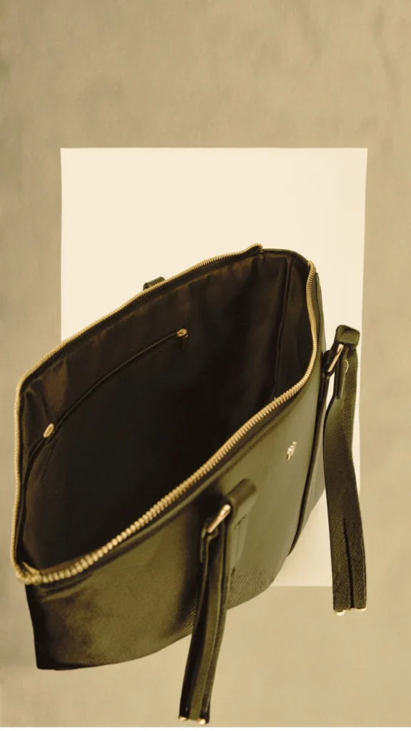 A high-angle view of a structured, open black pebble-grain leather tote bag set against a minimalist, warm neutral backdrop. The bag features an unzipped top opening that reveals a black fabric-lined interior with a small zippered pocket. It includes two top handles and a dangling leather strap detail accented with gold hardware.