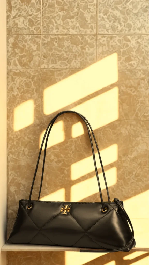 A sleek, black leather quilted East-West style handbag sits on a shelf against a warm neutral tiled wall. The bag features thin double shoulder straps, a zipper top closure, and a prominent gold Tory Burch logo on the front panel. Strong, geometric shadows from a window cast columns of bright afternoon sunlight across the background wall and the top of the purse.