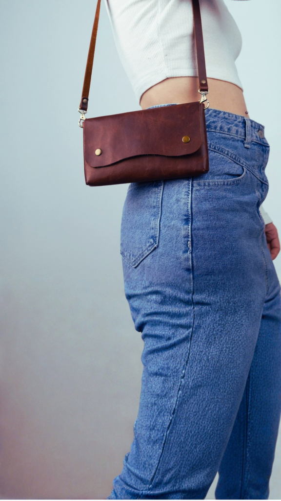 A side-profile shot of a woman wearing high-waisted blue jeans and a white ribbed crop top. A small, slim rectangular crossbody bag made of distressed brown leather rests at her hip. The handbag features a simple flap closure secured by two brass snap buttons and attaches to a thin leather strap with metallic silver lobster clasps.