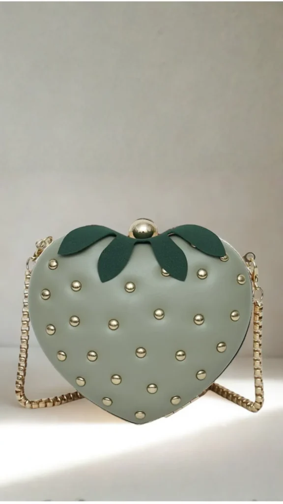 A whimsical, novelty crossbody handbag shaped like a strawberry from the file 3_9.png. The structured bag features a smooth, matte sage-green or light olive exterior studded with rows of round, polished gold rivets mimicking strawberry seeds. It is topped with four dark green faux-leather leaves that frame a metallic gold sphere clasp lock. A textured gold chain strap is attached to the sides, resting on a clean white surface against a minimalist cream background.