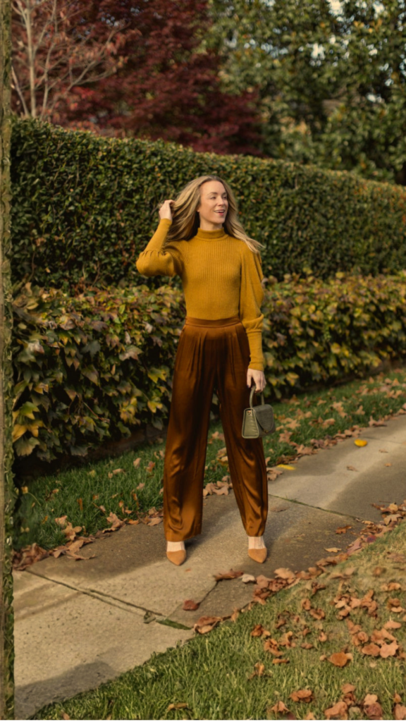 A blonde woman stands outdoors on a leaf-strewn neighborhood sidewalk with a dense green hedge and autumn trees behind her. She wears a warm mustard yellow mock-neck sweater featuring puff-shoulder detailing, paired with high-waisted, wide-leg satin trousers in a rich rust or copper brown hue. In one hand, she carries a structured olive green croc-embossed top-handle mini bag to complete her warm-toned fall look.