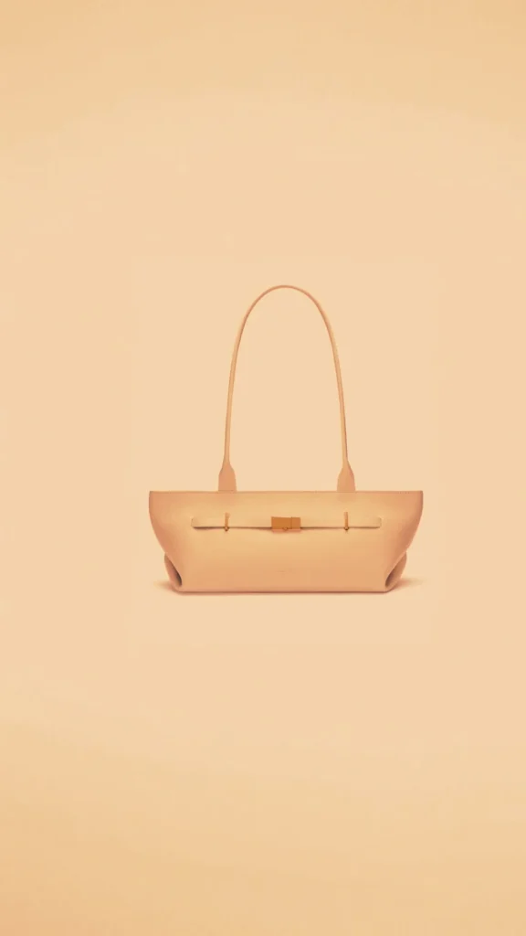 A minimalist, horizontal beige or light cream leather "east-west" shoulder bag centered against a solid cream background. The bag features clean lines, a single long, thin shoulder strap, and a subtle horizontal leather band across the front with minimal gold-toned hardware accents.