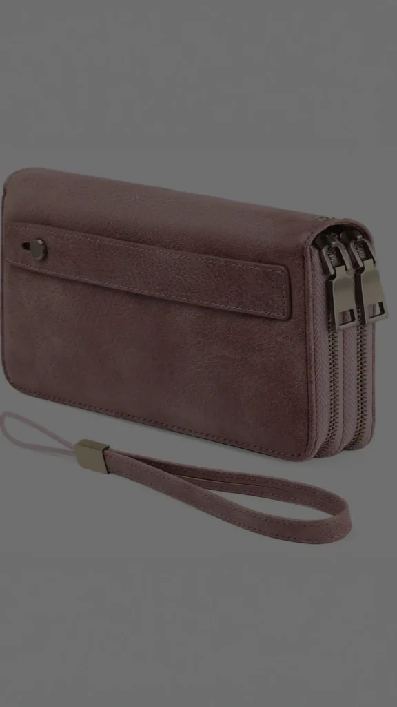 A structured dusty rose or mauve leather wristlet wallet shown from an angled side profile view against a solid grey background. The handbag features a textured, slightly weathered leather finish, a dual-zipper closure system with dark metallic pulls, and a matching leather wrist strap attached to one end. A flat horizontal strap with a small metal snap button is visible on the front face of the wallet.