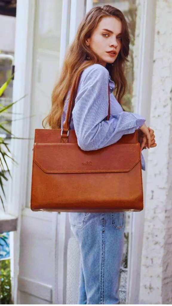 A young woman with long wavy hair looks back over her shoulder, wearing a striped blue button-down shirt and light-wash blue jeans. She carries a large, structured tan leather laptop tote bag or satchel under her arm. The sleek bag features clean lines, a front flap pocket with a subtle stamped logo, and long double shoulder straps.