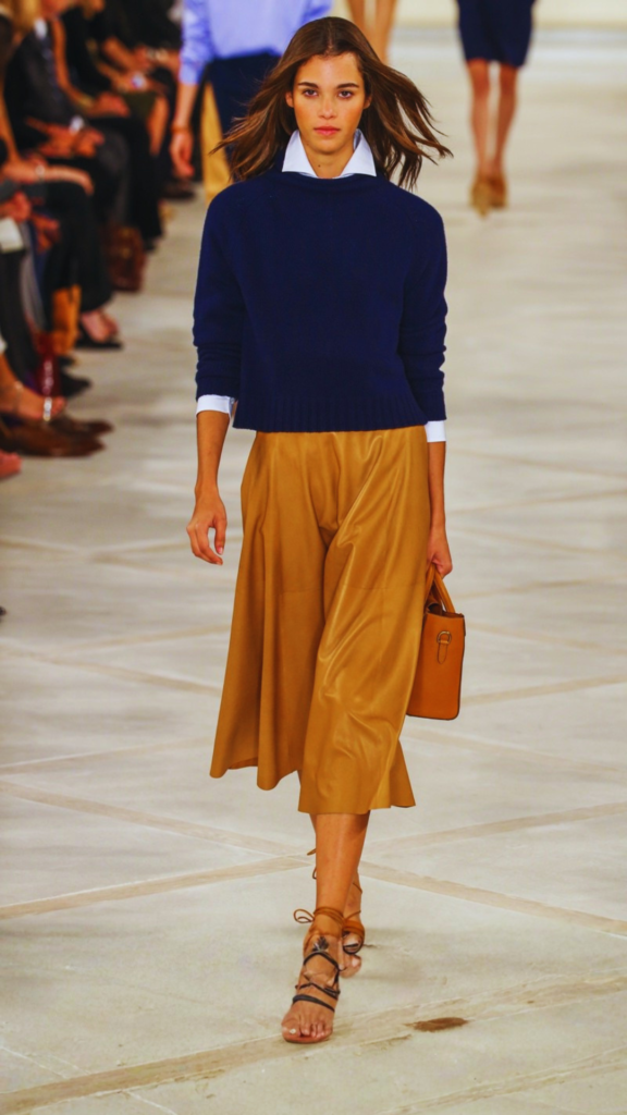 A fashion model walks down an indoor runway. She wears a navy blue crewneck sweater layered over a crisp white collared shirt, paired with a flowing, camel-colored midi skirt and strappy brown lace-up sandals. She carries a small, structured tan leather handbag in her left hand, completing a classic, polished aesthetic.