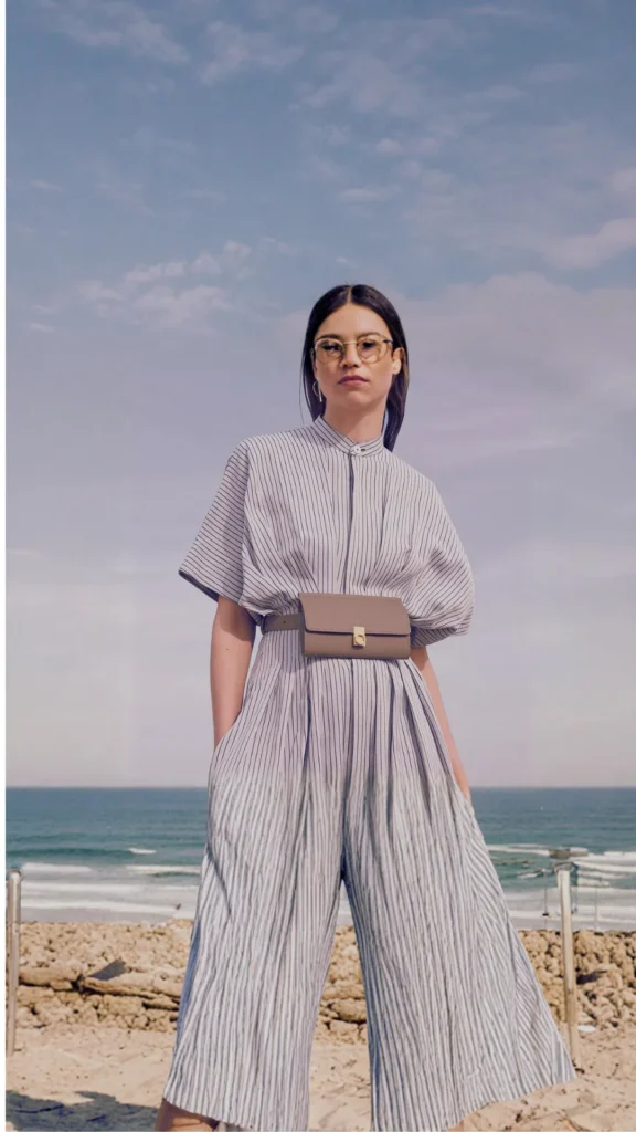 A woman standing outdoors against an ocean backdrop with a cloudy sky. She wears glasses and a modern, high-neck, short-sleeved jumpsuit with fine vertical stripes and voluminous wide-leg trousers. A structured, small taupe leather belt bag with a gold clasp is secured at her waist, defining the silhouette of the outfit.