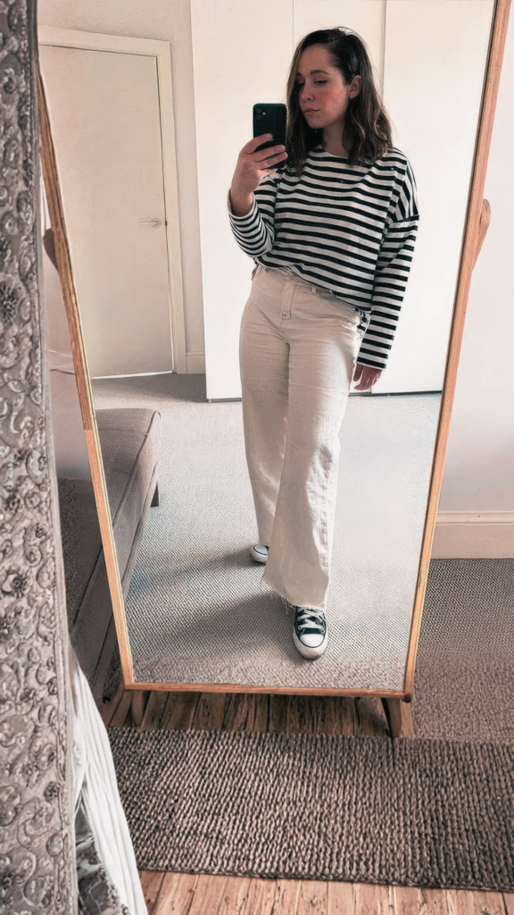 A young woman stands in a bright room with light-colored flooring and white walls, facing a large, decorative oval full-length floor mirror to take a selfie with her phone. She is dressed in a casual, minimalist style featuring an oversized cream-colored crewneck sweatshirt, light gray soft leggings, and clean white slip-on sneakers.