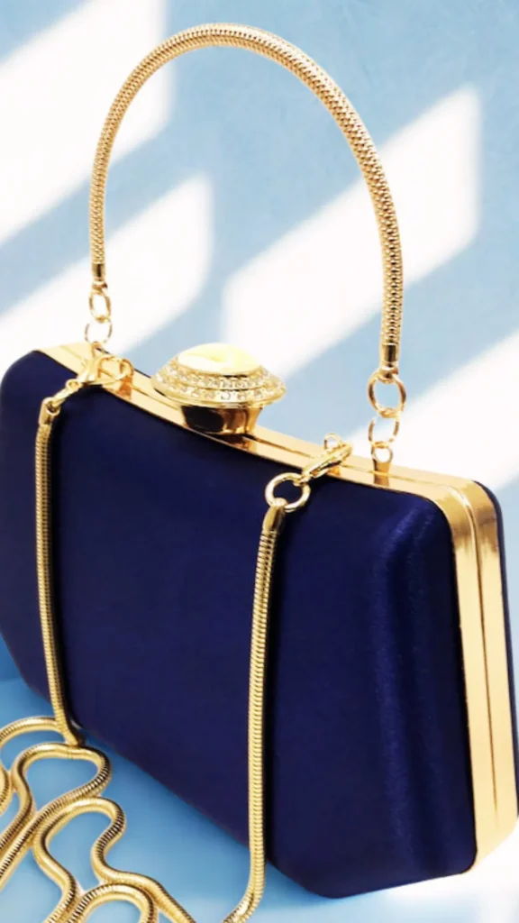 An elegant navy blue satin evening clutch bag displayed against a light blue backdrop with soft shadows. The rectangular, box-shaped handbag features a polished gold frame and a secure top clasp adorned with a large shimmering crystal element. It is equipped with a matching curved gold snake-chain top handle, and a longer gold chain strap pools gracefully on the surface next to the purse.