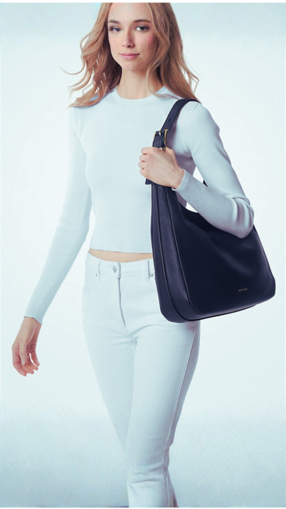 A blonde woman in a white long-sleeved crewneck top and matching white jeans walks forward against a clean white backdrop. She carries a large, structured navy blue or black pebble-grain leather hobo bag over her shoulder. The minimalist handbag features a wide shoulder strap with a prominent gold buckle accent near the top.