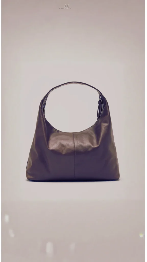 A large, slouchy dark brown or espresso-colored leather hobo bag centered against a solid, light neutral background. The minimalist handbag features a soft, crescent-shaped silhouette with a wide single shoulder strap integrated seamlessly into the body, a central vertical seam detail, and a subtle zipper closure at the top.