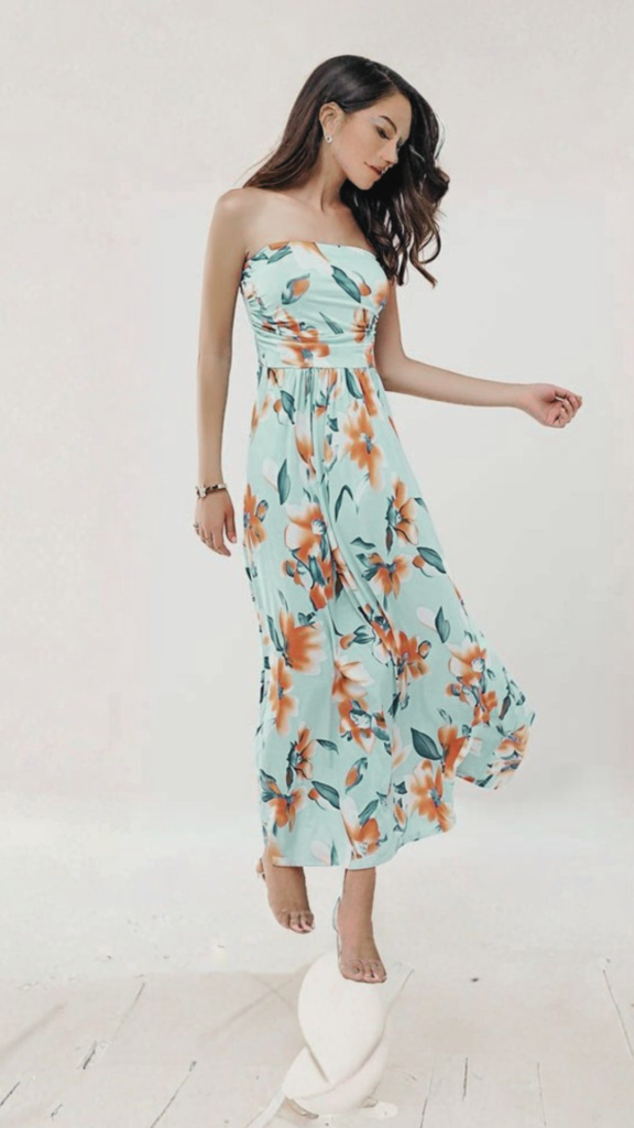 A young woman stands outdoors on a sunny day next to a leafy tree, smiling over her shoulder. She is wearing a sleeveless maxi dress with a blue and white floral pattern, a wide-brim straw sun hat with a frayed edge, and small gold hoop earrings. She carries a spacious, medium-wash denim tote handbag with thick shoulder straps over her arm.