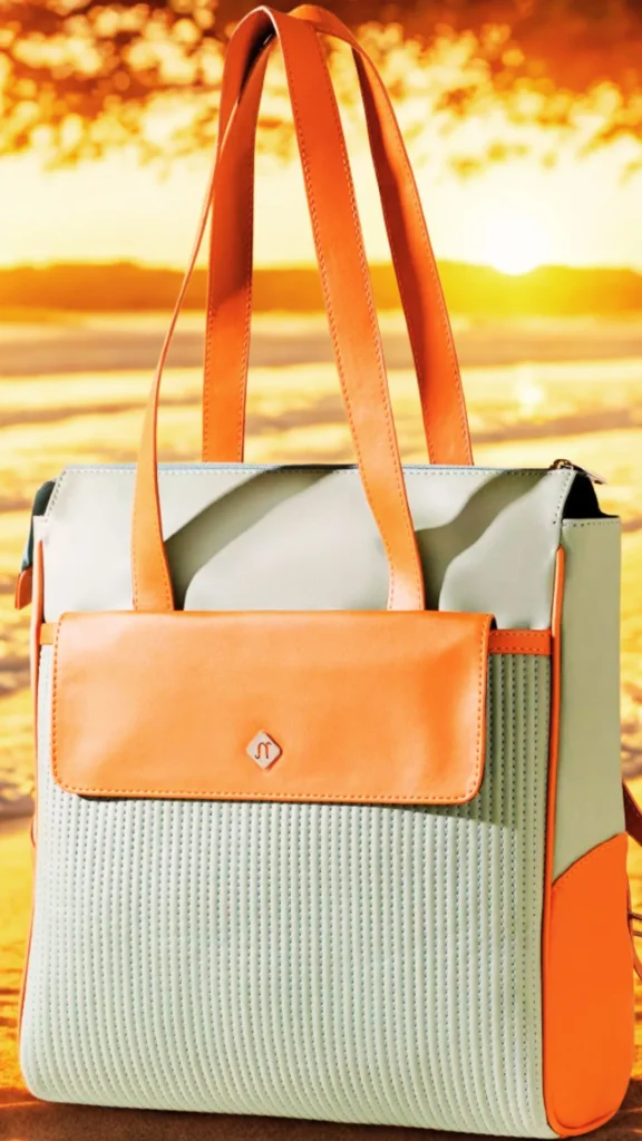 A structured, convertible backpack tote handbag stands on a sandy beach against a blurred golden sunset over the ocean. The bag features a light mint green or pale sage colored body with fine vertical quilting on the front pocket panel. It is accented with vibrant orange faux-leather trim, dual top shoulder handles, side panels, a front flap pocket closure, and a small silver diamond-shaped metal logo emblem.