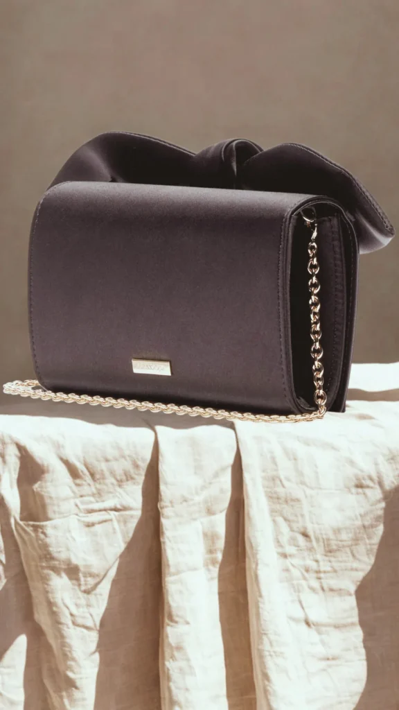 A structured rectangular evening clutch bag made of smooth, dark charcoal gray or black satin fabric, resting on a draped beige linen cloth. The purse features a delicate, interwoven gold chain shoulder strap across the front, a small rectangular gold logo plate on the lower flap, and a soft, gathered top accent creating a stylish, knotted bow effect.