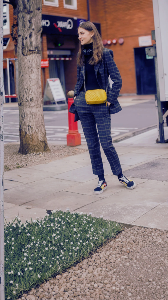 A young woman stands on a city sidewalk wearing a dark plaid trouser suit over a black turtleneck, paired with patterned sneakers and headphones around her neck. She has her hands in her pockets and wears a small, vibrant yellow quilted rectangular crossbody bag with a silver chain strap.
