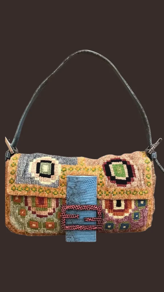 A product shot from 1 (1)_2.webp showcasing a vintage beaded Fendi Baguette handbag against a dark brown background. The bag features an intricate, colorful beaded pattern in shades of orange, green, blue, and black, resembling needlepoint embroidery. It is uniquely accented with a bright blue lizard-skin patterned leather front strap and a contrasting pink lizard-skin patterned "FF" buckle closure. A thin, dark blue leather shoulder strap completes this iconic shoulder bag.