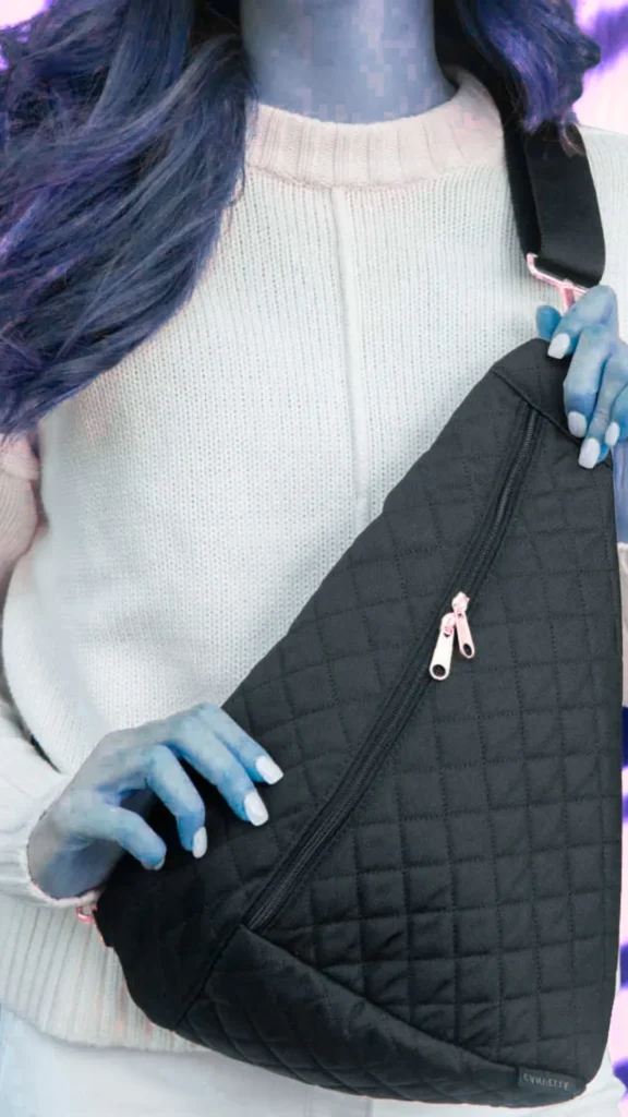 A close-up shot from the file 1_9.webp shows a person wearing a textured, cream-white knit sweater and a black quilted sling bag draped across their chest. The tear-drop shaped bag features a prominent diagonal zipper with silver metal pulls and a wide black fabric strap resting on the shoulder. The person's hair is a vibrant blue-purple shade, and their skin has a distinct pale blue monochromatic overlay tint. Their hands, featuring white painted nails, rest lightly on the bag and its strap.
