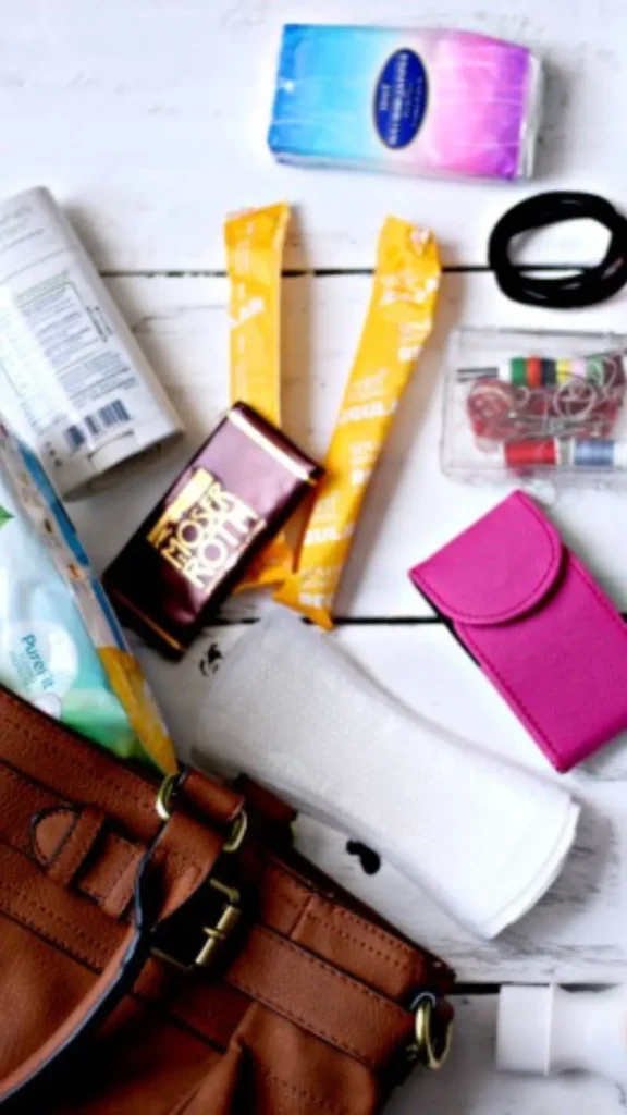 An image from file 1 (1)_5.webp showing a flat-lay view of everyday essentials spilled out from a brown faux-leather tote bag onto a white wooden surface. The contents include a blue and pink gradient package of tissues, two yellow snack bars, a dark red Moser Roth chocolate bar, a small clear sewing kit, a black hair tie, a bright pink compact case, a roll of white paper towels or wipes, and various hygiene products.