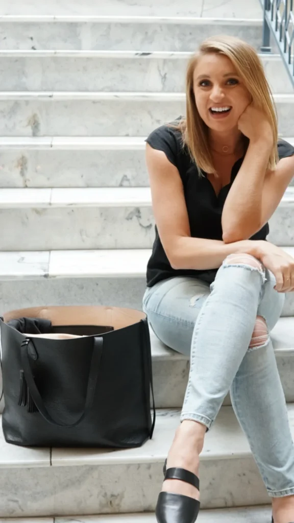 An image from file 1 (1)_7.webp featuring a blonde woman smiling cheerfully while sitting cross-legged on light gray marble indoor stairs. She is wearing a sleeveless black top, ripped light-wash blue jeans, and black leather mule flats. Next to her on the step sits a large, structured black leather tote bag with dual shoulder straps and a decorative black leather tassel hanging from one handle. The wide bag is open, revealing a spacious, light tan-colored interior lining.