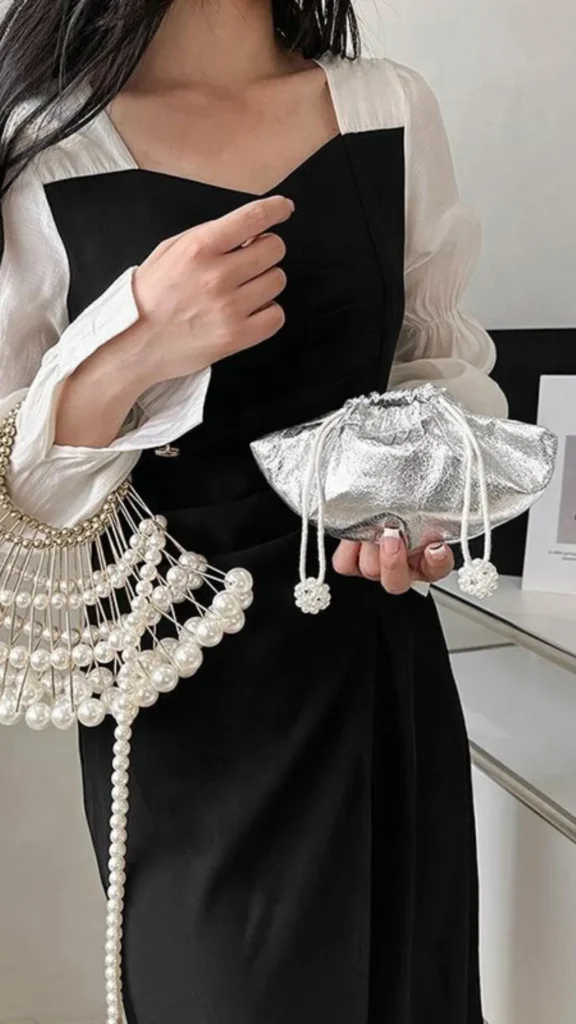 A close-up style photograph from 1.webp showcasing unique, statement-making fashion accessories. A person wearing a black dress over a white puff-sleeve blouse holds a small, metallic silver foil drawstring pouch pouch bag in one hand. Dangling from her other arm is an intricate, architectural fan-shaped bag constructed completely from strings of large, glossy white pearls that form a sculptural cage design.
