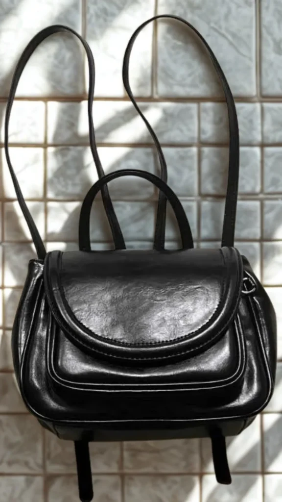A flat-lay lifestyle shot from 1_3.webp showcasing a minimalist black leather mini backpack resting on a white-tiled background under soft sunlight and leaf shadows. The unbranded bag features a glossy, textured leather finish, a rounded front flap closure with a matching curved piping detail, a rigid top handle, and extended thin leather backpack shoulder straps, embodying a clean and structured aesthetic.