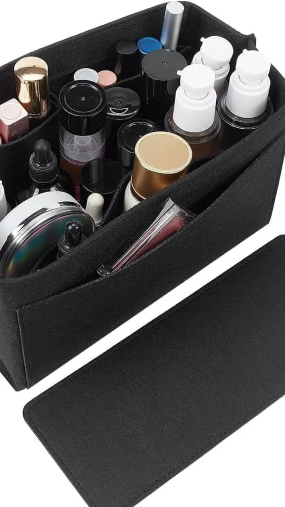 A high-angle product shot from 1_6.webp showing a rectangular black felt handbag organizer insert neatly filled with cosmetics and skincare products. The main compartment is partitioned into smaller sections holding various items, including pump bottles, dropper bottles, lipsticks, and compacts with metallic caps. An empty matching black felt base liner or divider rests flat on the white surface directly in front of the main pouch.