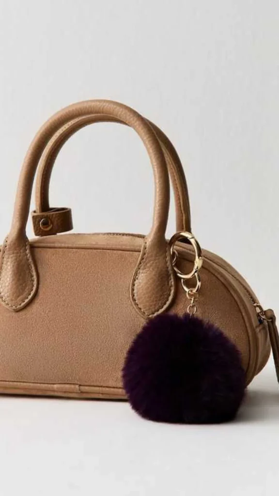 A close-up product photograph from 1_7.webp showcasing a tan suede bowler-style handbag decorated with a fluffy bag charm. The small neutral-colored bag features curved leather top handles, matching stitched trim, and a top zipper. Attached to the front handle base with a gold-toned circular spring clasp is a large, dark purple faux-fur pom-pom charm, adding a distinct contrast in color and texture against a light grey background.