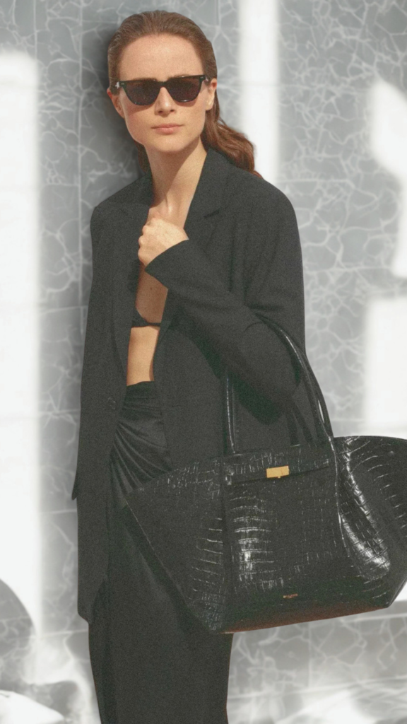 A three-quarter fashion photograph from the file 1.png features a woman posing outdoors in a monochromatic black ensemble. She wears an open black blazer over a matching bralette, a draped black skirt, and dark sunglasses. Over her forearm rests a very large, structured black tote bag with a striking glossy croc-embossed texture and a subtle gold clasp mechanism. The background consists of a textured light grey wall reflecting strong, angular sunlight and shadows.