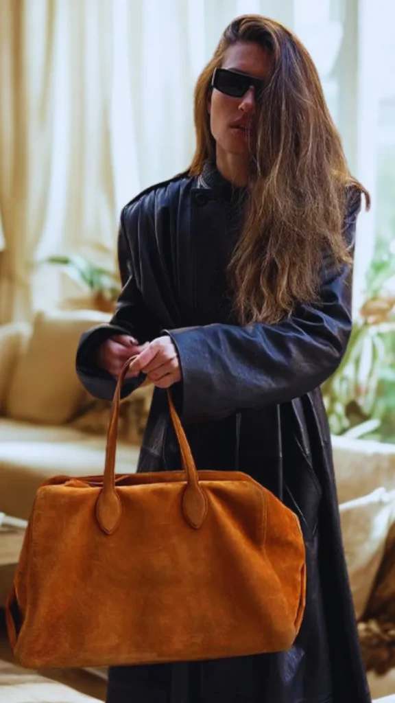 A medium shot from the file 1_12.png shows a person with long, flowing brown hair wearing oversized black sunglasses and a long, dark leather trench coat. The individual is holding a large, structured tan or camel-colored suede tote bag by its twin leather top handles. The background is a brightly lit, soft-focus indoor living space featuring a cream-colored couch, drapes, and indoor plants.