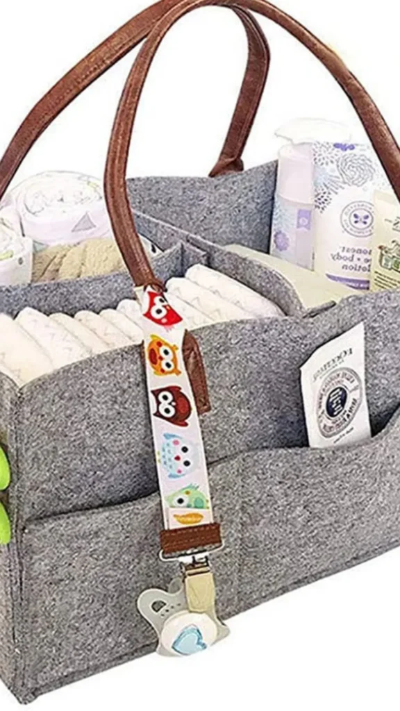 A close-up product photograph from 1_4.jpg showcasing a grey felt baby diaper caddy organizer basket packed with baby essentials. The structured organizer features multiple internal compartments neatly filled with a row of folded white diapers, baby lotions, wipes, and a tube of cream tucked into an external side pocket. It has brown leather-look top handles, and a white pacifier clip strap decorated with colorful cartoon owls hangs from the handle base, holding a baby pacifier.