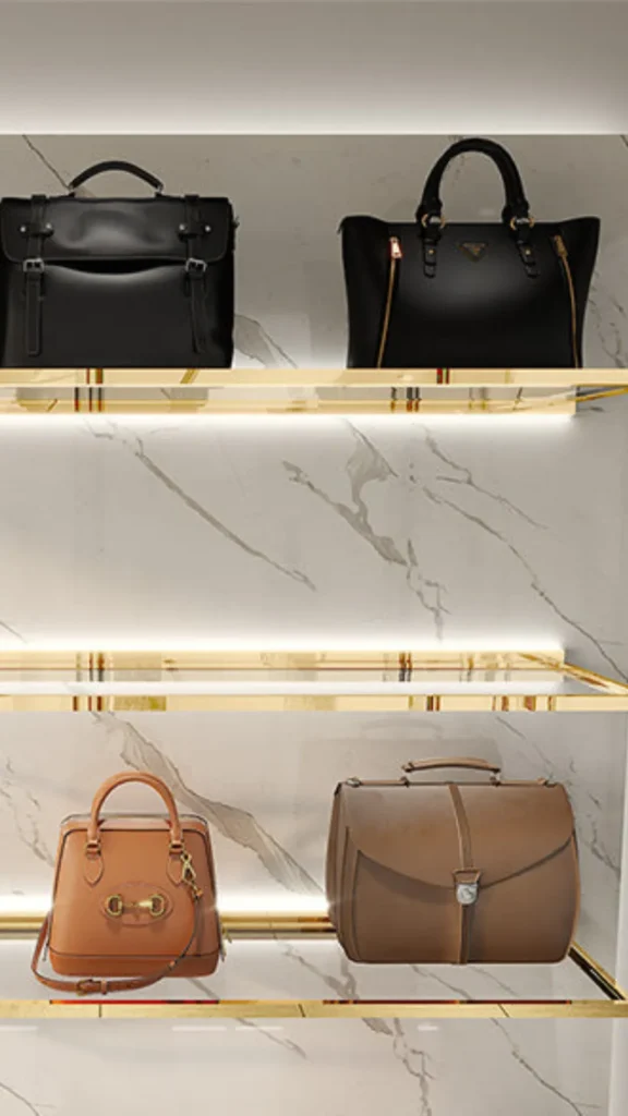 An image from file "1_16.webp" showing a luxurious vertical display of designer handbags arranged on floating shelves. The shelves feature polished gold frames with built-in LED strip lighting that illuminates each level against a white marble wall backdrop. The top shelf displays two large black bags: a structured briefcase style with double buckles and a classic tote with gold zippers. The lower shelf showcases two tan bags: a smaller structured handbag with a top handle and shoulder strap, and a larger rounded doctor-style satchel bag.