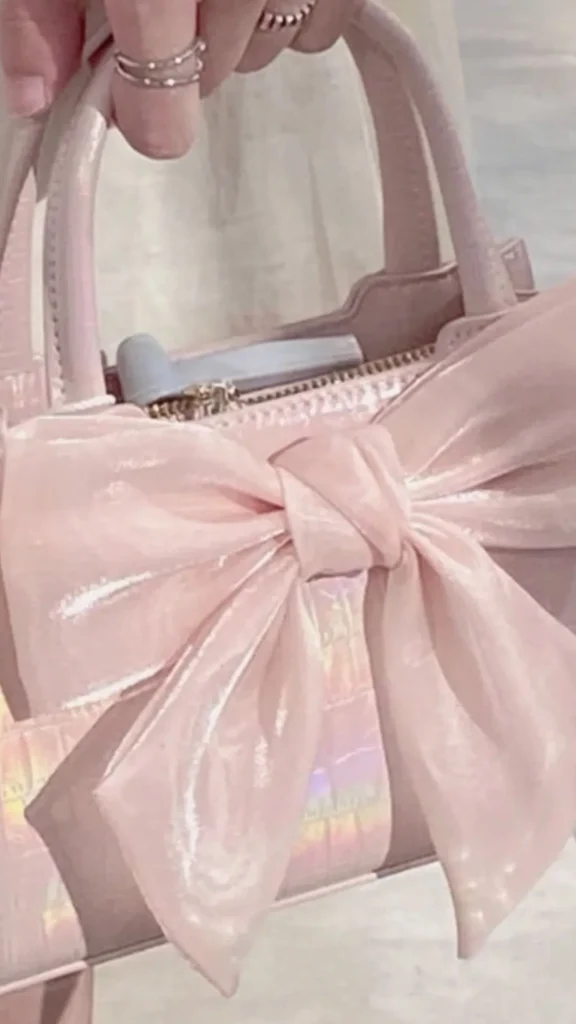 An image from file "1_17.webp" showcasing a close-up view of an ultra-feminine, coquette-style handbag. A hand adorned with delicate silver rings holds the top handle of the mini bag. The handbag features a glossy, iridescent light pink croc-embossed texture that reflects subtle rainbow hues. A large, prominent satin or organza fabric bow in a matching soft blush pink shade is tied beautifully across the front of the bag, capturing a romantic and playful balletcore aesthetic.