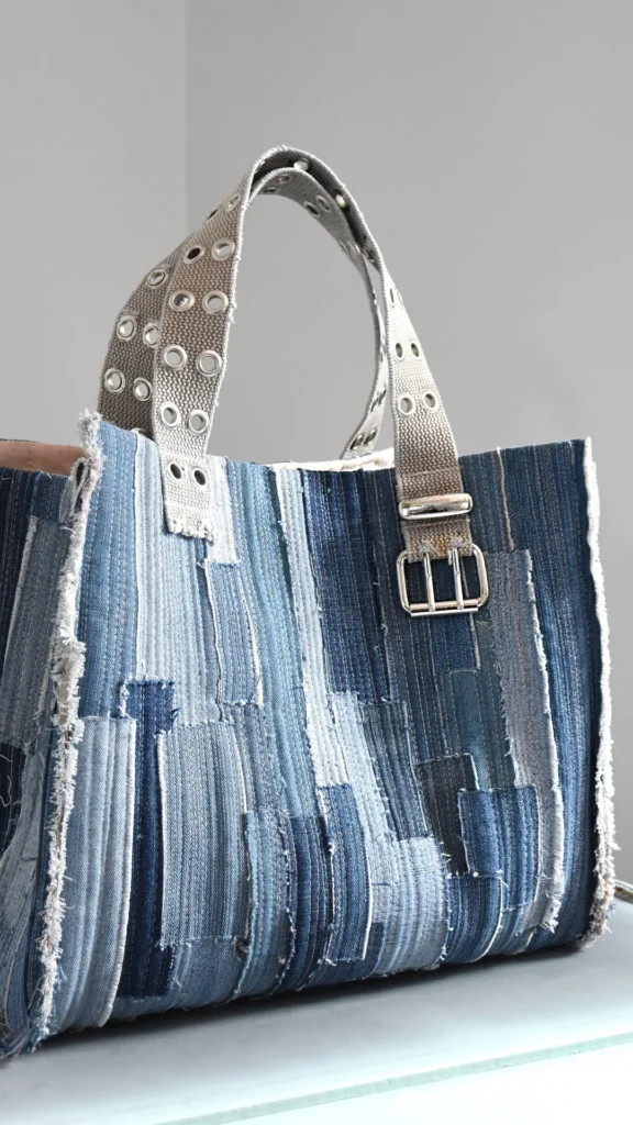 An image from file "1_12.jpg" showing a vertical product shot of a rectangular tote bag handmade from upcycled denim fabric. The main body of the bag features a patchwork style, stitched together using vertical strips of denim in varying shades of blue, from light wash to dark indigo, with raw, frayed edges along the seams and upper border. It has two top handles made from thick, grey woven straps heavily decorated with shiny silver metal grommets. A prominent silver metal buckle is attached to the base of one of the handles on the front right side, reinforcing its rugged, reconstructed design.