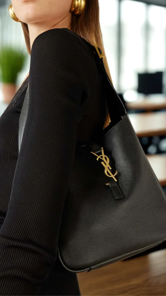 An image from file "1_19.webp" showcasing a close-up side view of a person wearing a black ribbed long-sleeve top and carrying a black Saint Laurent Le 5 à 7 hobo bag tucked under their arm. The bag is crafted from supple, textured black grained leather and features an integrated leather shoulder strap along with the iconic gold metal Cassandre YSL monogram hook closure securely fastened across the top opening. The person's face is partially cropped, revealing large, chunky gold hoop earrings. The blurred background shows a modern office or cafe setting with wooden tables and indoor greenery.