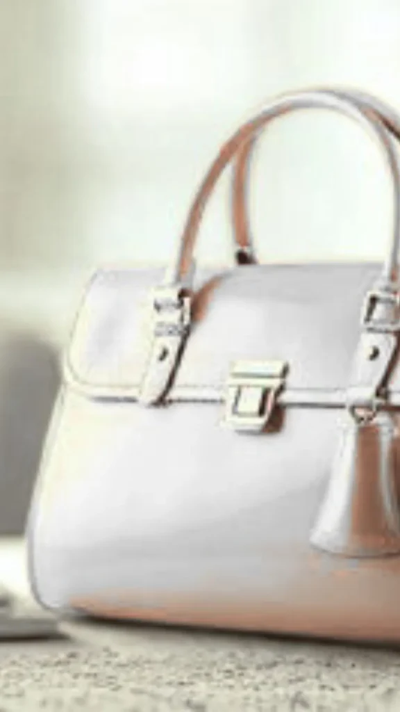 A close-up shot from the file 1_10.webp displays a structured handbag made of a glossy, reflective light-grey or silver patent leather material. The bag sits on a textured surface and features two arched top handles, a front flap secured with a silver clasp closure, and a matching leather clochette tag hanging from the front handle base. Soft, bright light illuminates the side and front of the purse, catching its metallic sheen against a blurred background.