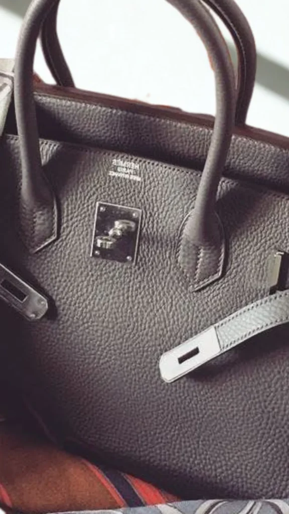 A close-up product photograph from the file 1.webp showcases a classic luxury tote handbag in dark charcoal or deep taupe pebbled grain leather. The bag features structured rolled top handles and iconic silver hardware detailing, including a central turn-lock closure plate and a leather strap pulled through a silver bracket. The silver embossed logo stamp "HERMÈS PARIS / MADE IN FRANCE" is visible just below the upper rim.