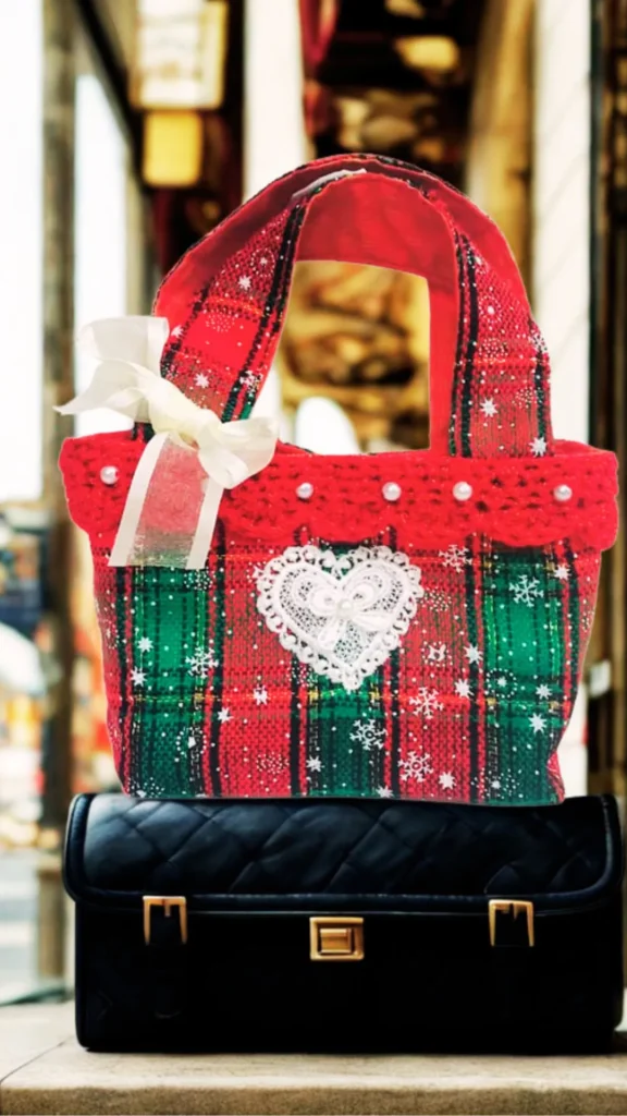 A close-up shot from the file 1_8.webp displays a handmade festive tote bag balanced on top of a black quilted leather briefcase. The handmade bag features a red and green plaid fabric patterned with tiny white snowflakes. Its top rim is bordered by a textured red ruffled trim adorned with small white pearls, and a large white lace heart is stitched into the center of the front panel. A decorative white satin ribbon bow is tied to the base of one of the thick fabric top handles. The black bag underneath features a gold twist-lock closure flanked by two gold buckles, all set against a softly blurred background.
