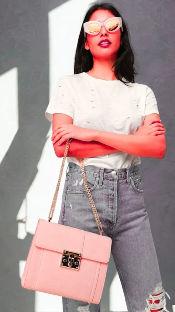 A portrait from the file 1.webp shows a person standing with crossed arms, wearing a distressed white t-shirt, light gray ripped jeans, and pink-framed sunglasses. They carry a structured pastel rose pink leather shoulder bag with a gold chain strap draped over their arm. The handbag features a prominent square gold turn-lock closure on the front flap. The image is styled with a distinct warm, high-contrast pink-toned overlay filter against a dark background.