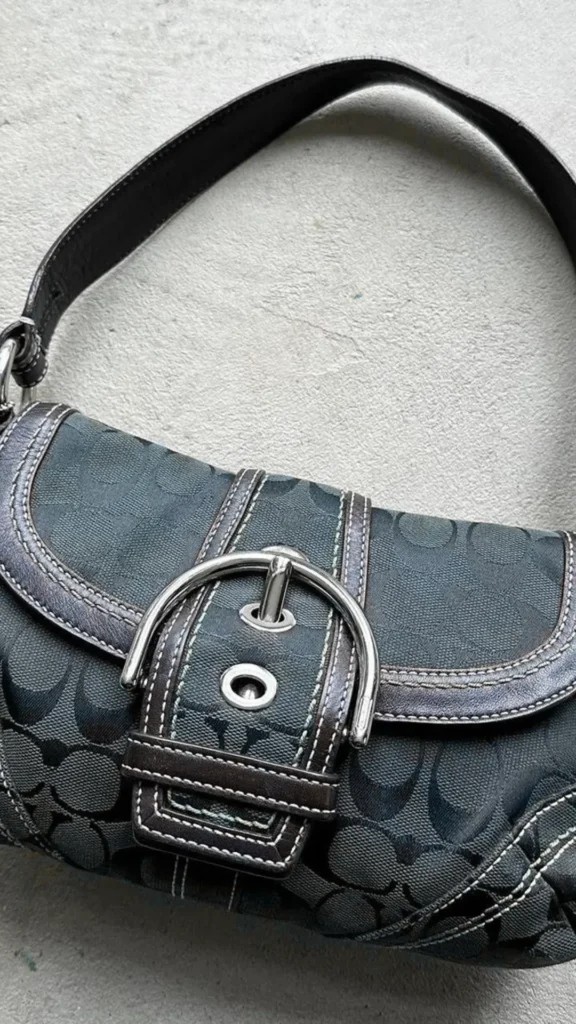 A close-up product photograph from the file 1.jpg features a vintage-style Coach baguette shoulder bag resting on a textured gray concrete surface. The handbag is made of dark charcoal or black canvas material woven with the signature Coach "C" monogram pattern. It features a dark leather shoulder strap, detailed white contrast stitching along the edges, and a prominent front flap secured by a thick leather strap with a large, polished silver-metal horseshoe buckle closure.