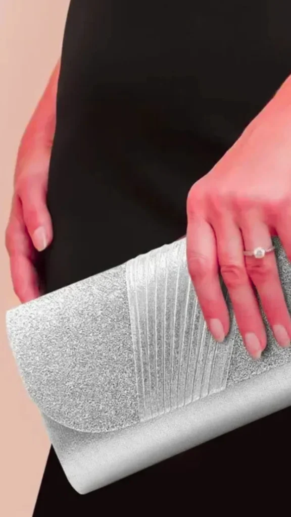 A close-up photograph from the file 2 (1).webp shows a person wearing a classic black dress holding a shimmering silver evening clutch bag. The clutch features a fine glitter texture with a decorative, vertically pleated silver satin band running down the center of its front flap. The person’s right hand rests elegantly on top of the purse, showcasing a delicate ring with a clear gemstone on the ring finger against a soft pink background.