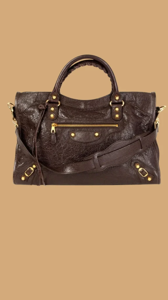 A product shot from 2 (1)_2.webp showcasing an iconic Balenciaga City bag against a warm tan background. The handbag is crafted from dark brown, highly textured, crinkled agneau leather that has a signature "lived-in" finish. It features classic gold-tone metal studs and buckles, a zippered front pocket with a long leather tassel pull, hand-stitched braided top handles, and a detachable leather shoulder strap with a padded section.