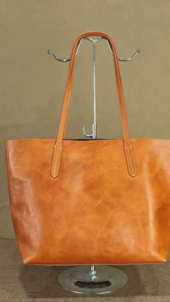 An image from file 2 (1)_4.webp showing a front view of a classic tan leather tote bag displayed on a silver metallic table stand. The spacious, structured bag features a textured, warm caramel-brown leather finish and two long, slender shoulder straps attached to the front panel with clean stitching. The display stands against a neutral, plain brown backdrop.