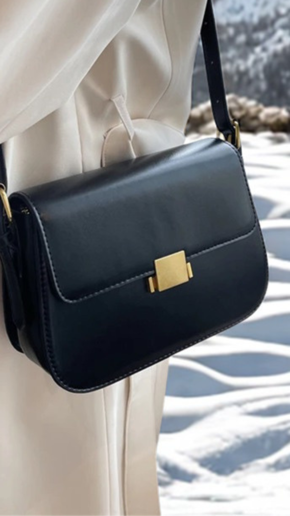 A close-up fashion photograph from the file 2.png shows a person's torso as they model a sleek, black leather structured handbag worn over the shoulder. The small, rectangular purse features a front flap closure with a unique, minimalist silver metallic geometric lock. The person is dressed in a crisp, white long-sleeved button-up shirt neatly tucked into a pair of high-waisted, vibrant cobalt blue denim jeans, creating a clean, modern color contrast.