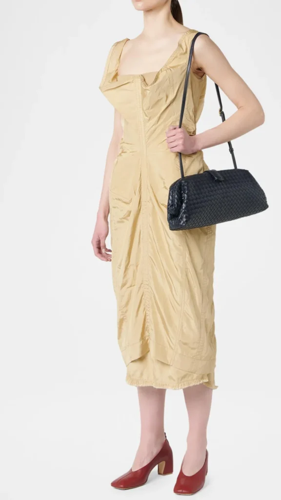 A full-length fashion shot from 2.webp of a model standing against a white background. She is wearing a sleeveless, crinkled beige or light gold midi dress with structured draping and dark red square-toe low heels. Over her left shoulder, she carries an elongated, dark navy blue or black Bottega Veneta Lauren 1979 style clutch bag featuring a distinct Intrecciato woven leather pattern, a frame closure, and a thin matching shoulder strap.